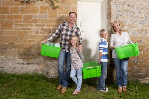 A family of four, including a man, a woman, and two children, stands outdoors against a textured stone wall, each holding bright green plastic rubbish bins. The man is positioned on the left, smiling and wearing a checked shirt and jeans, with his bin held in his right hand, resting on the ground. The young girl, standing in front of him, is smiling and dressed in casual clothes, holding her bin close to her body. To the right, the boy, wearing a blue and white striped shirt and jeans, is holding a green bin and looking toward the woman, who is positioned on the far right, wearing a white blouse and jeans, also holding a green bin. The ground is covered in short green grass, suggesting a garden or yard setting. The scene appears to be part of a rubbish removal or garden clearance activity, aligning with services offered by Garden Clearance Harlesden in the local London area, possibly near the NW10 postcode. Natural light illuminates the scene, giving a clear view of the family's casual clothing and the vibrant colour of the bins, emphasizing a community-based approach to waste disposal or garden clearance services.