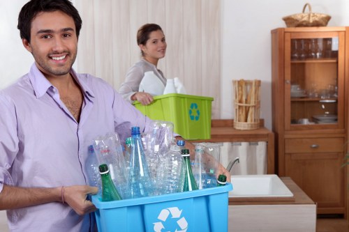 A man with dark hair and a light purple shirt is smiling and holding a blue recycling bin filled with empty plastic bottles in a domestic kitchen setting. The bottles are clear with green and blue labels, some with caps on, and appear to be squeezed or flattened. In the background, a woman with brown hair tied back is standing near a green recycling box placed on a wooden kitchen counter, which is empty in its interior. The kitchen features light-colored curtains, a wooden cabinet with glass doors containing dishes, and a white sink. The scene is well-lit, emphasizing the cleanliness and organization of the space, which is typical for a household preparing to dispose of recyclable rubbish related to waste management services such as those provided by Garden Clearance Harlesden. This image highlights domestic recycling activities within a typical kitchen environment.