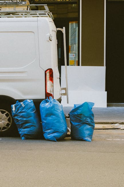 A concrete wall with a red metal door covered in graffiti tags and a 'Keep Clear' sticker, surrounded by black rubbish bags and discarded cardboard and packaging materials piled at the base of the doo