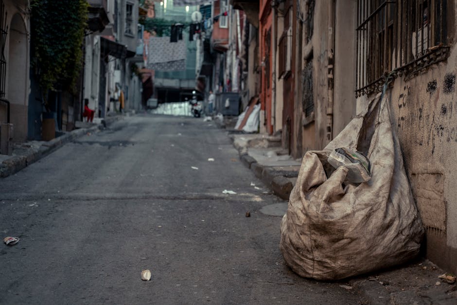 A middle-aged man with dark hair, wearing a black T-shirt with white graphic text, is leaning forward and using a wooden stick to place a large white sack filled with rubbish into a stainless steel li